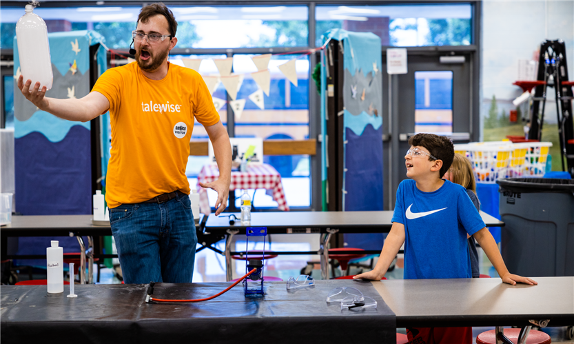 A man in an orange shirt performs a science experiment with a student volunteer